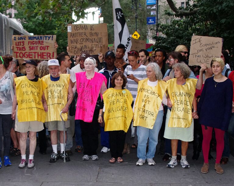 Eine Gruppe älterer Menschen steht in einer größeren Menschenmenge während einer Demonstration. Mehrere Leute tragen gelbe Westen mit der Aufschrift "Granny Peace Brigade". Andere halten Protestschilder in der Hand.